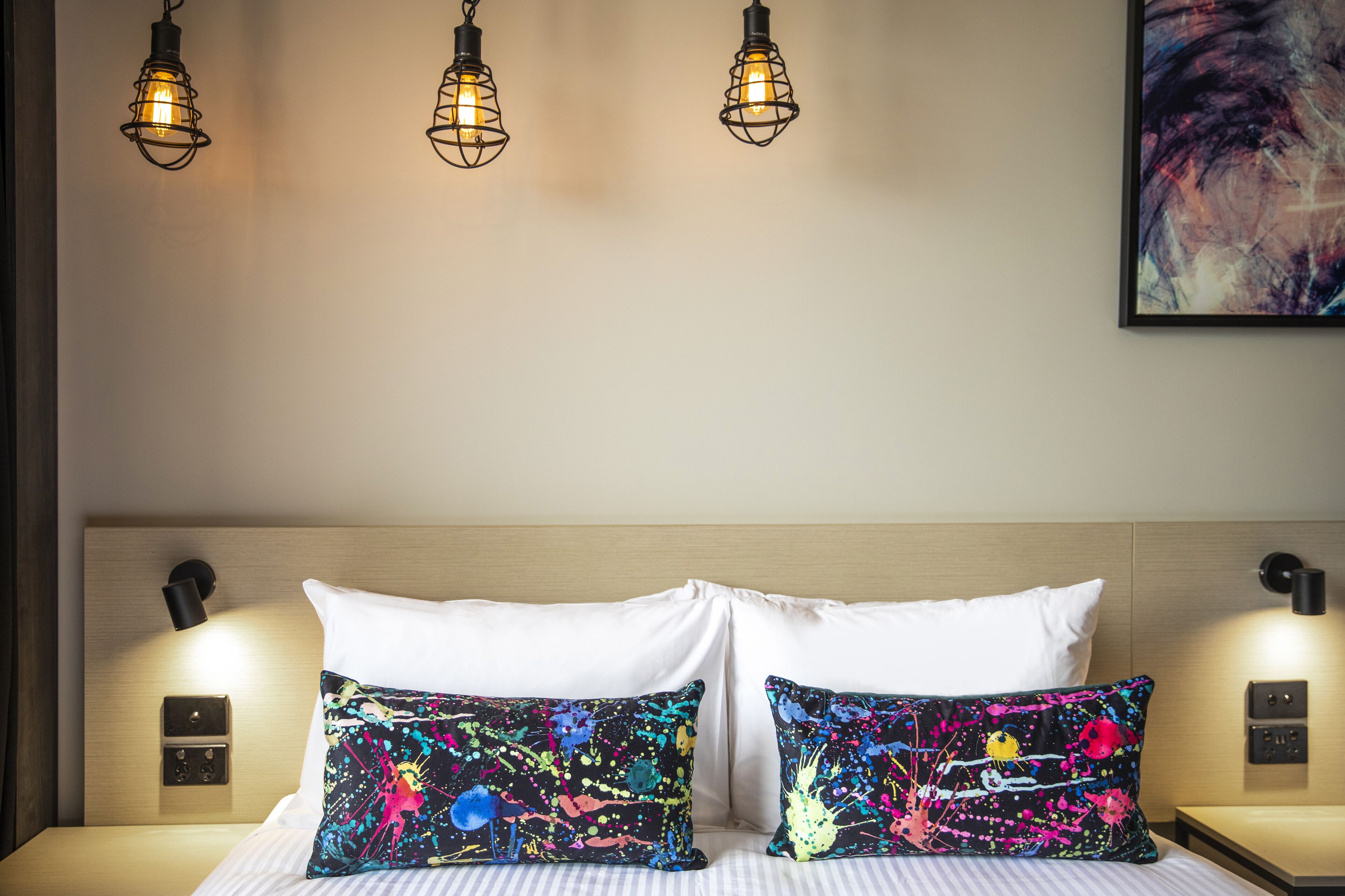 Quarto de hotel com uma cama, duas almofadas brancas, uma almofada colorida com padrão abstrato e três luminárias pendentes.