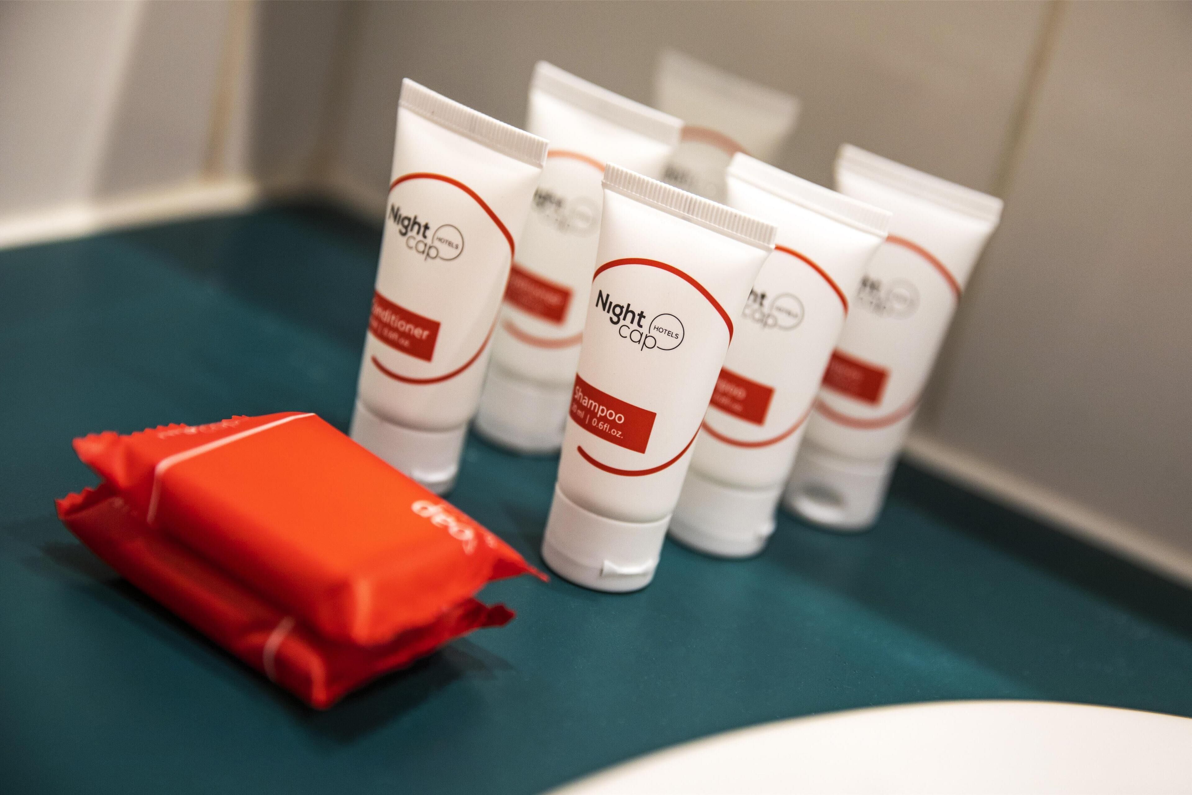 Une rangée de produits de toilette d’hôtel blancs, avec des inscriptions rouges et noires sur une surface turquoise.
