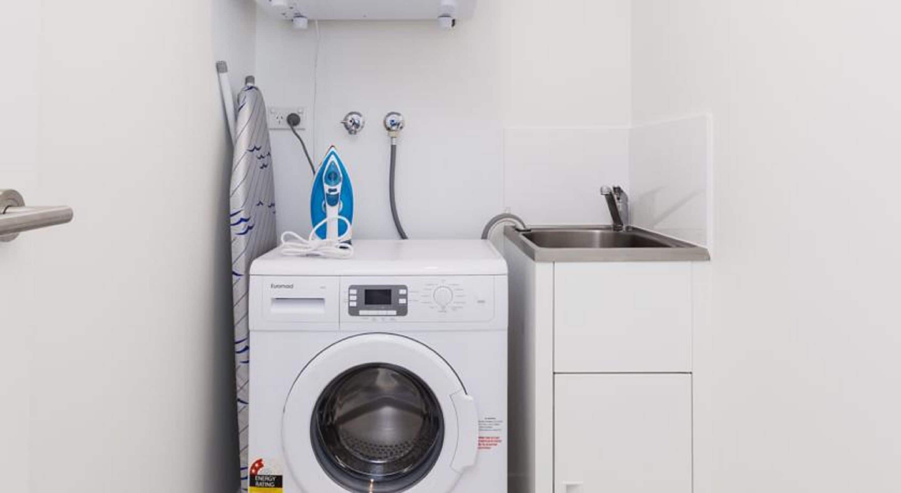 Une petite buanderie équipée d’un lave-linge à chargement frontal, d’un évier et d’un porte-serviettes.