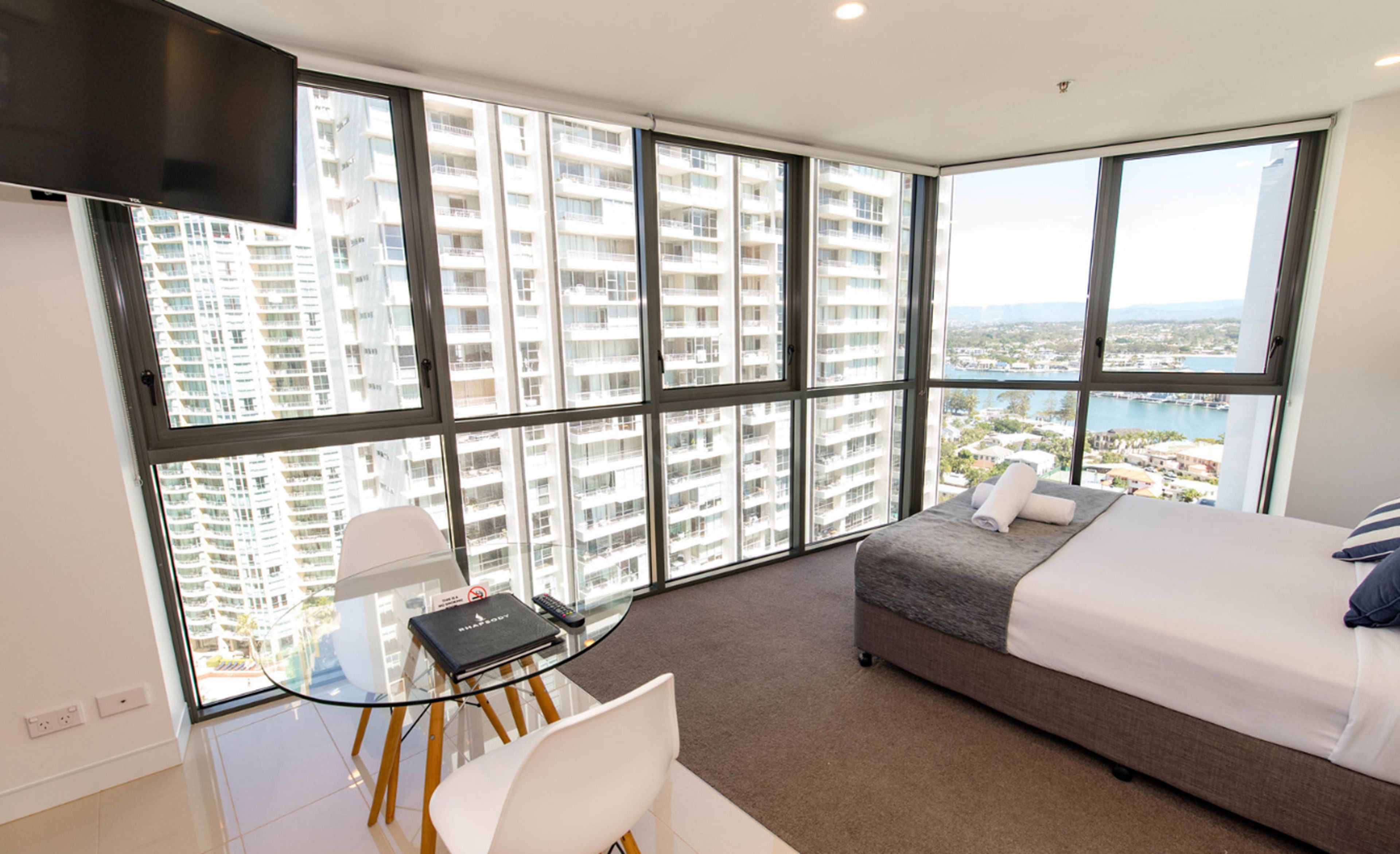 A modern hotel room with a large window, a bed, a desk with a chair, and a view of high-rise buildings and a body of water.