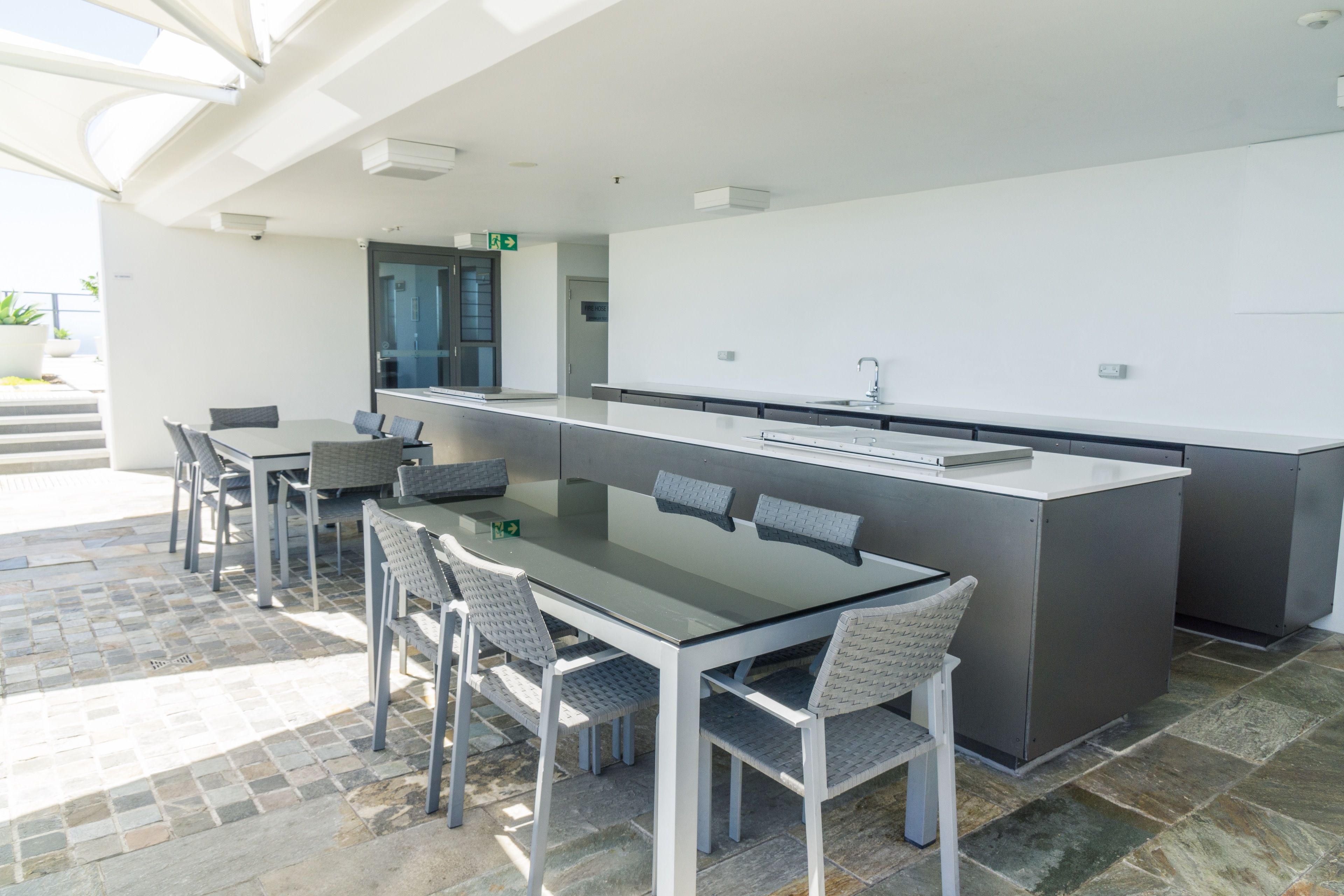 Un área de comedor exterior moderna con una mesa de vidrio y sillas blancas, una encimera de cocina y un piso de baldosas de piedra.