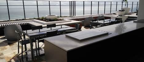 An outdoor terrace with a view of the ocean, featuring a dining area with tables and chairs, a seating area with cushions, and potted plants.