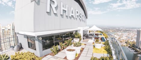 A modern building with the name 'Rhapsody' prominently displayed. It features a rooftop terrace with potted plants and seating areas.