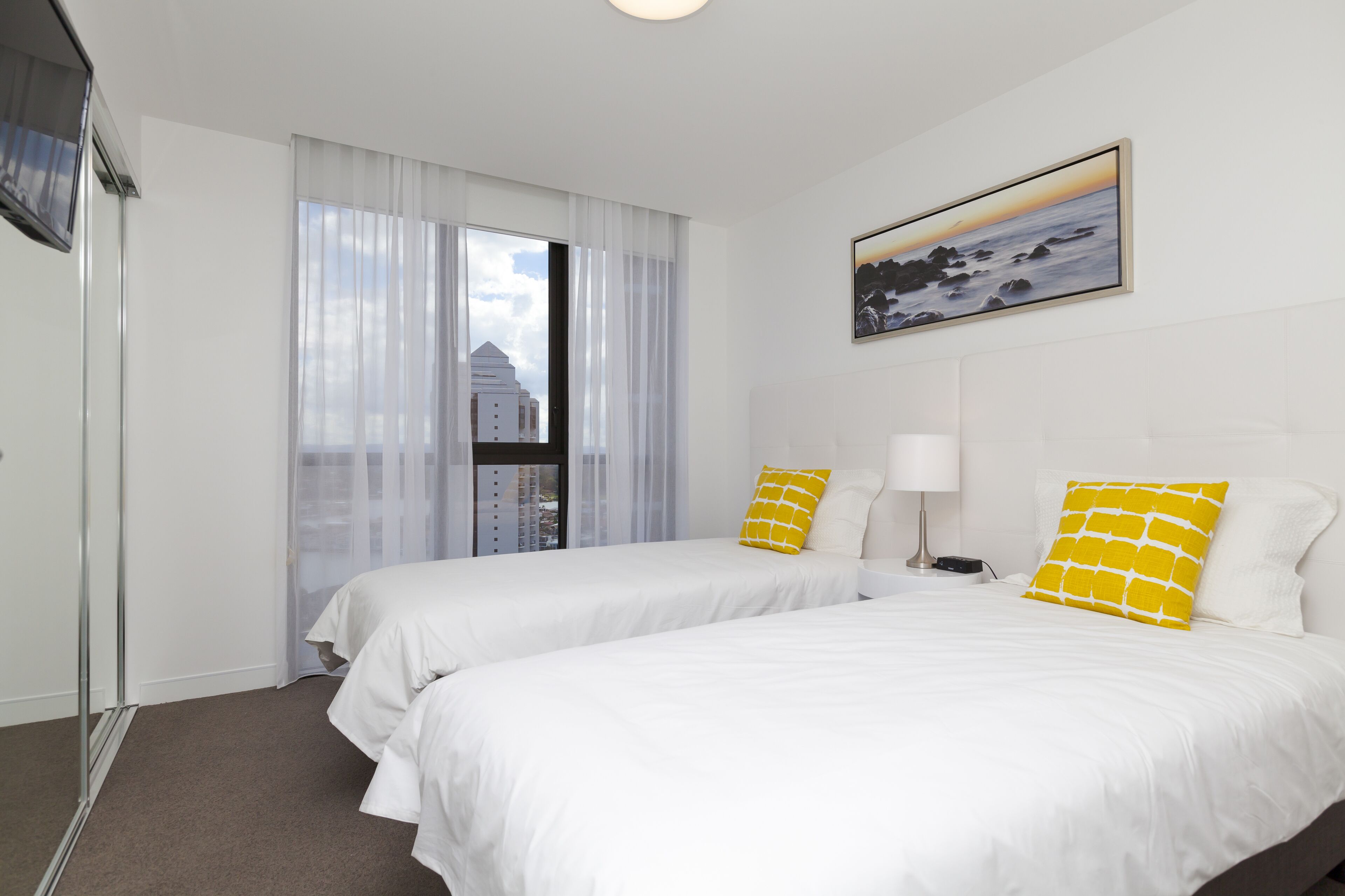 A hotel room with two beds, a window with curtains, a painting on the wall, and a nightstand with a lamp.