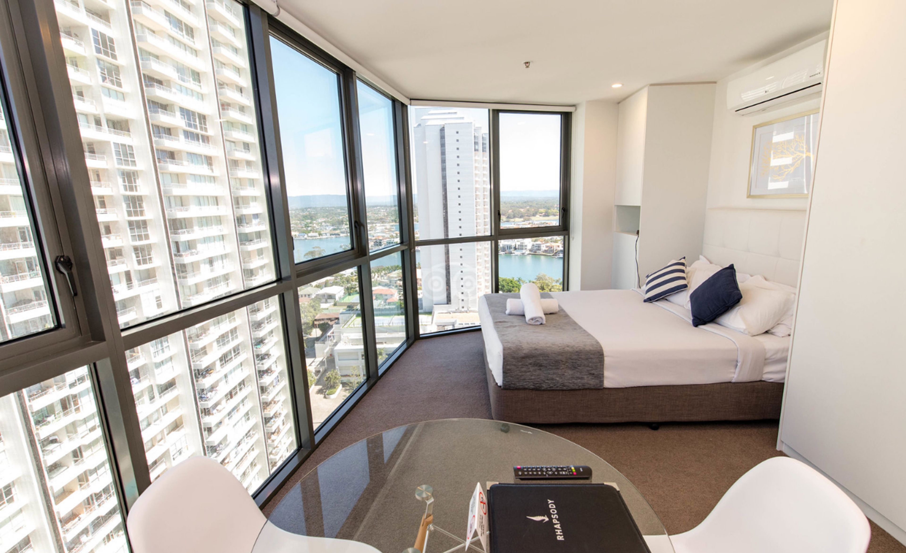 A modern hotel room with a large bed, a round glass table, and a view of high-rise buildings.