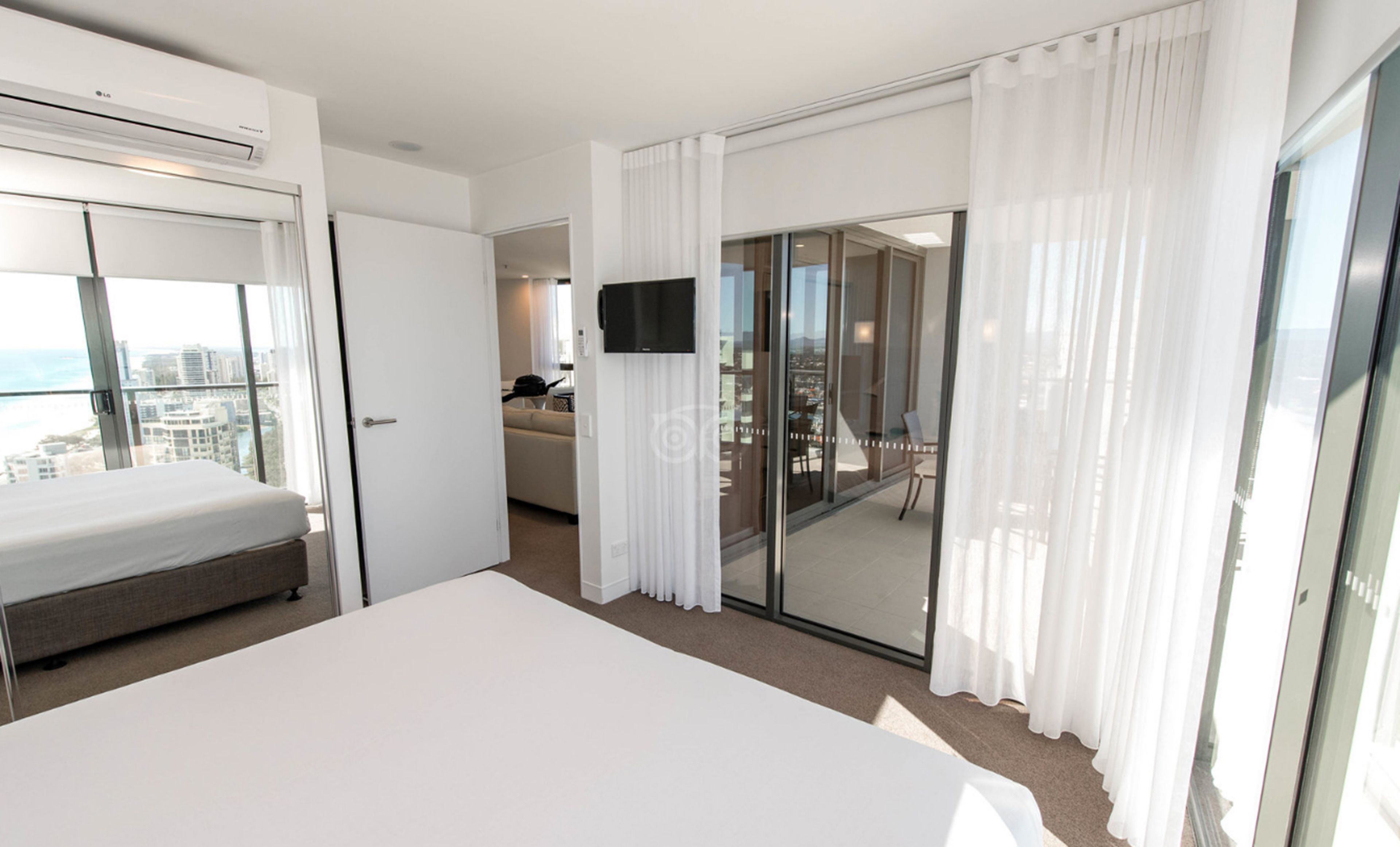 A modern hotel room with a large bed, a TV, a balcony with a city view, and a sliding glass door.