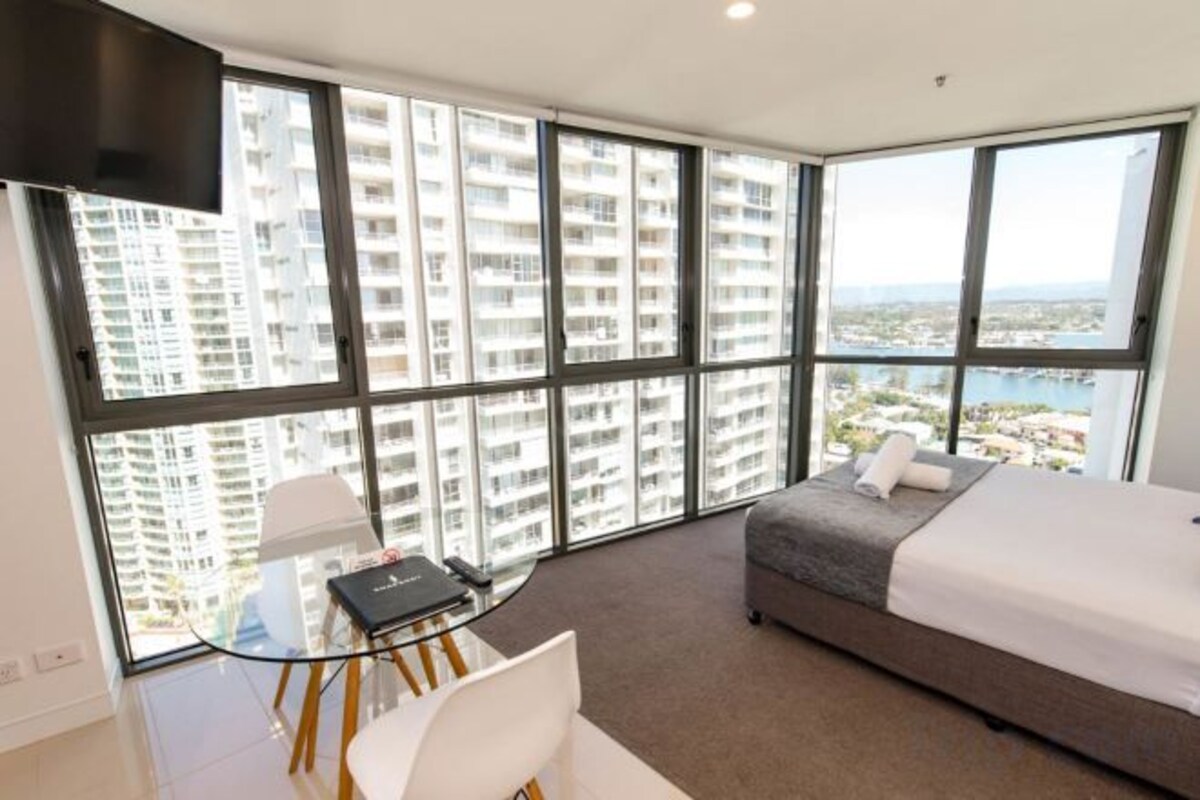 A hotel room with a large window, a bed, a glass table, and a chair.