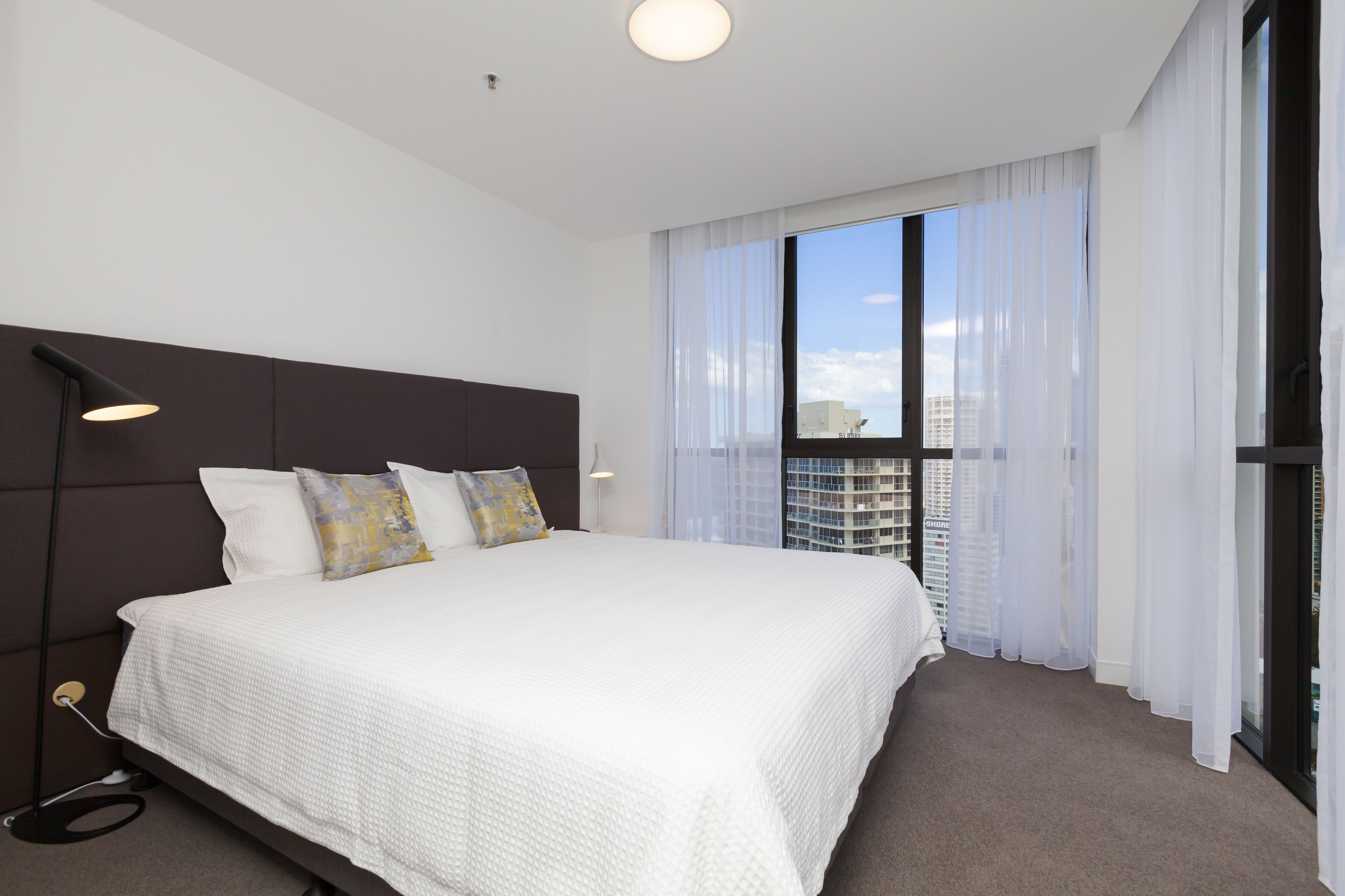 A modern hotel room with a large bed, a headboard, a window with sheer curtains, and a view of the cityscape.