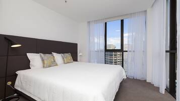 A modern hotel room with a large bed, a headboard, a window with sheer curtains, and a view of the cityscape.