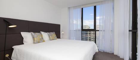 A modern hotel room with a large bed, a headboard, a window with sheer curtains, and a view of the cityscape.