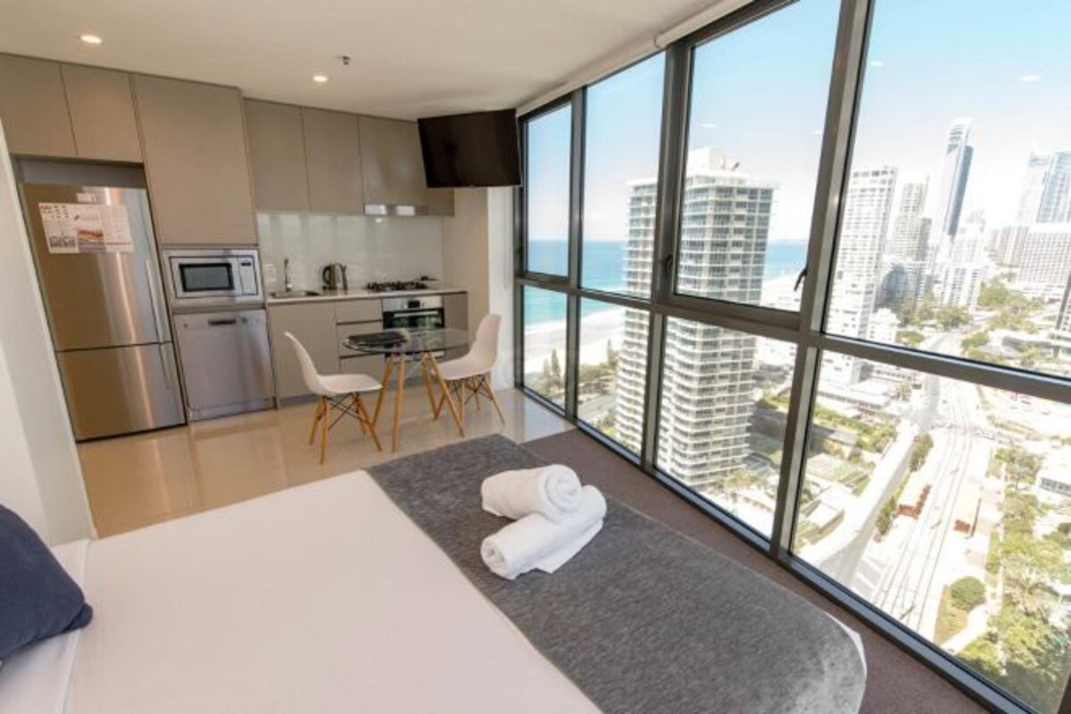 Une chambre d’hôtel moderne dotée d’un grand lit, d’une petite kitchenette, d’un coin repas et offrant une vue sur la ville grâce à de grandes fenêtres.