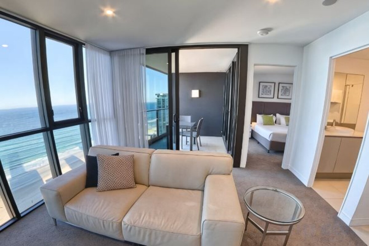 A modern hotel room with a balcony, a sofa, a dining table, and a kitchen area.