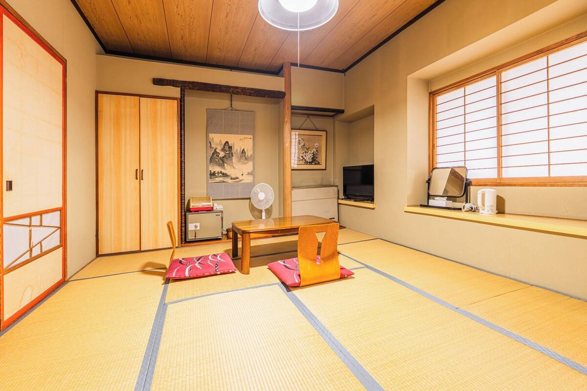 A traditional Japanese room with tatami flooring, a wooden ceiling, a sliding door, a television, a fan, and a window with shoji screens.