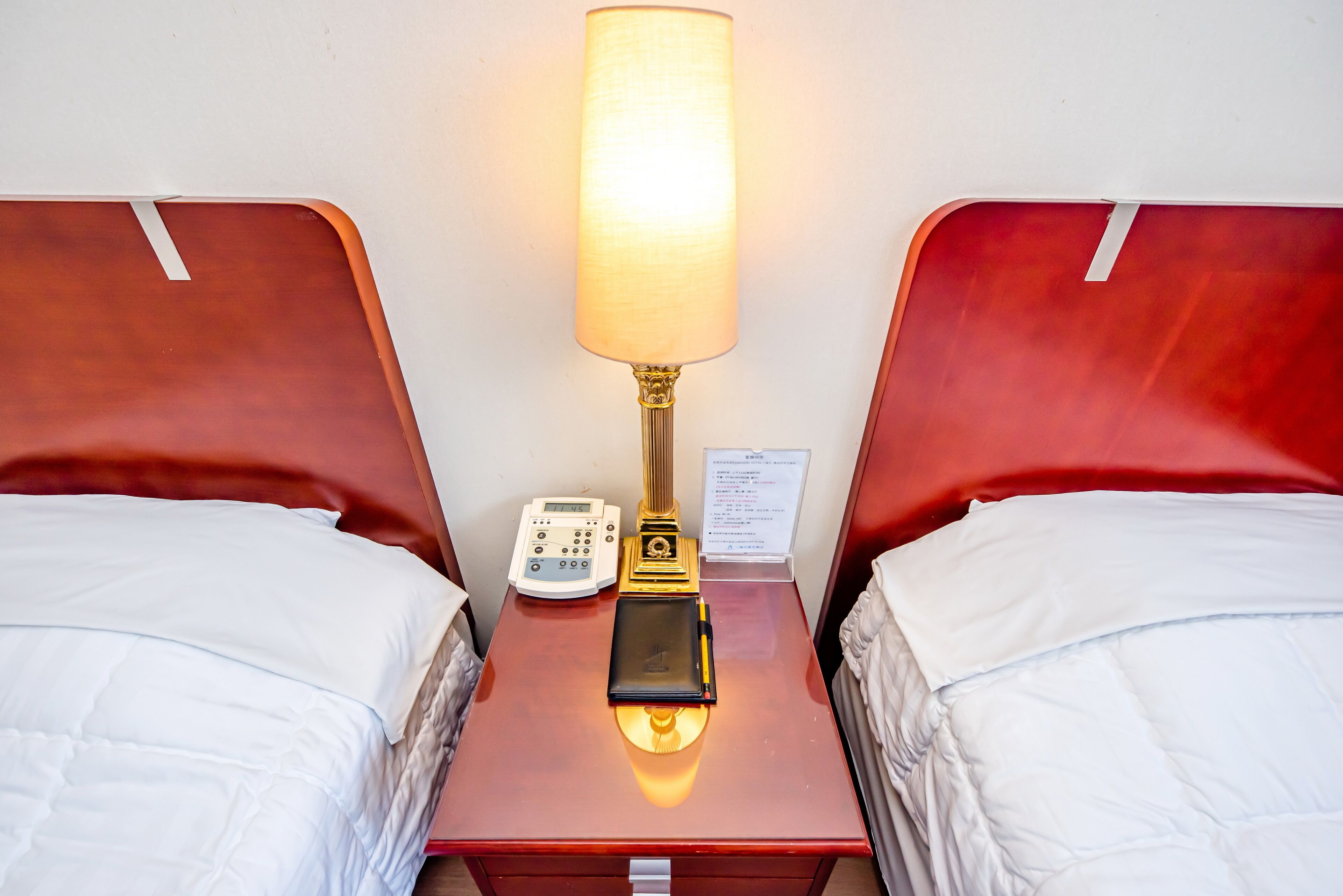 Habitación de hotel con dos camas, una mesita de noche con lámpara, un teléfono y una tarjeta pequeña.