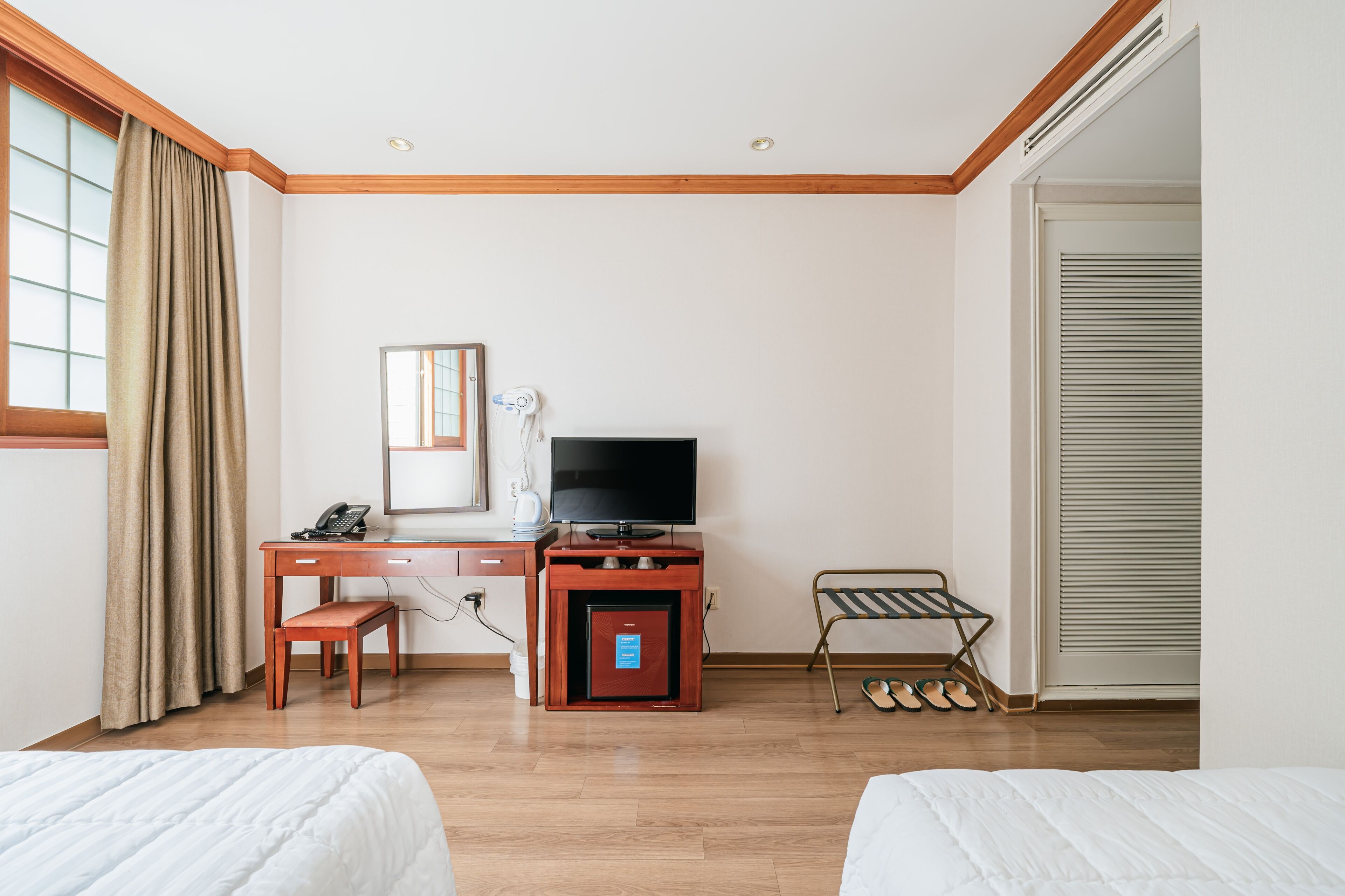 Hotellihuone, jossa on kaksi sänkyä, työpöytä, jossa on televisio, tallelokero ja kenkäteline.