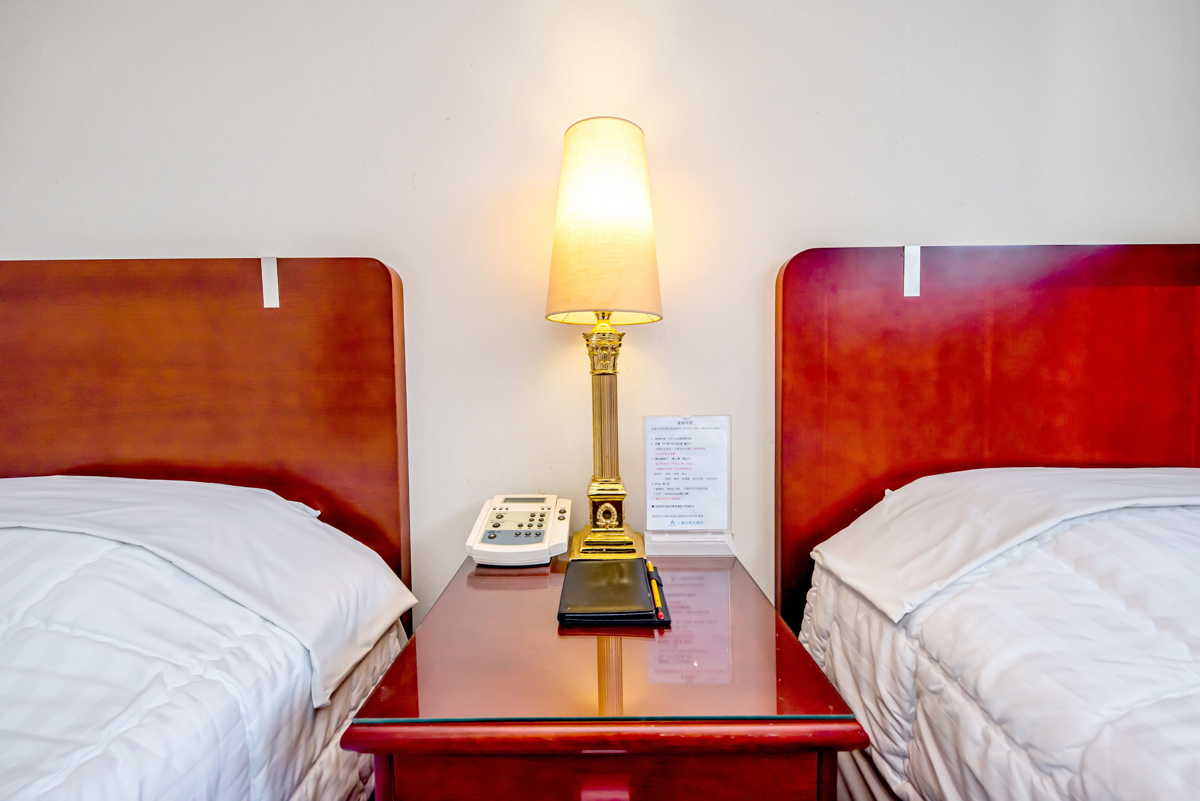 A hotel room with two beds, a bedside table with a lamp, a phone, and a menu on the wall.