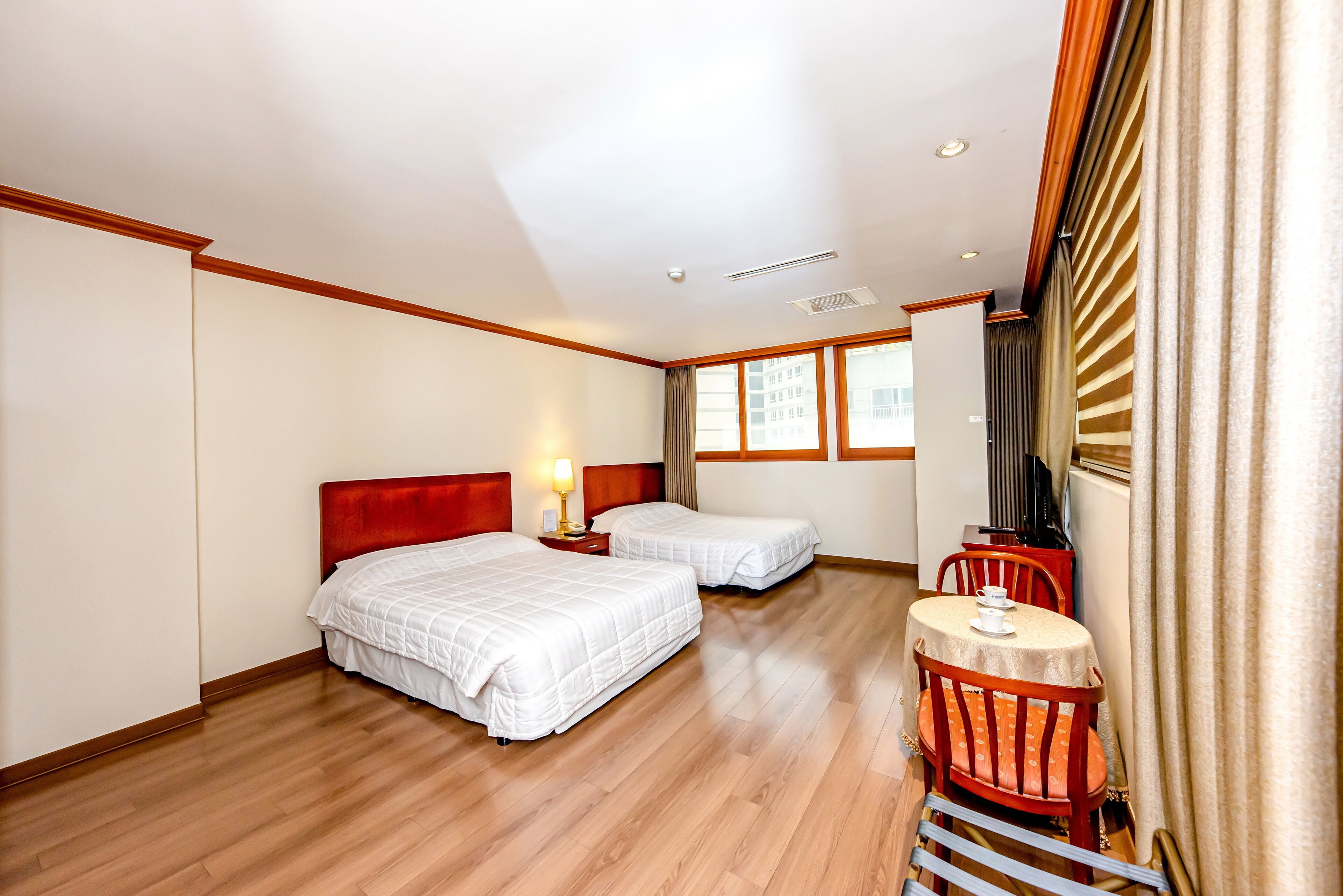 Una habitación de hotel con dos camas, una mesita con una silla y un ventanal con cortinas.