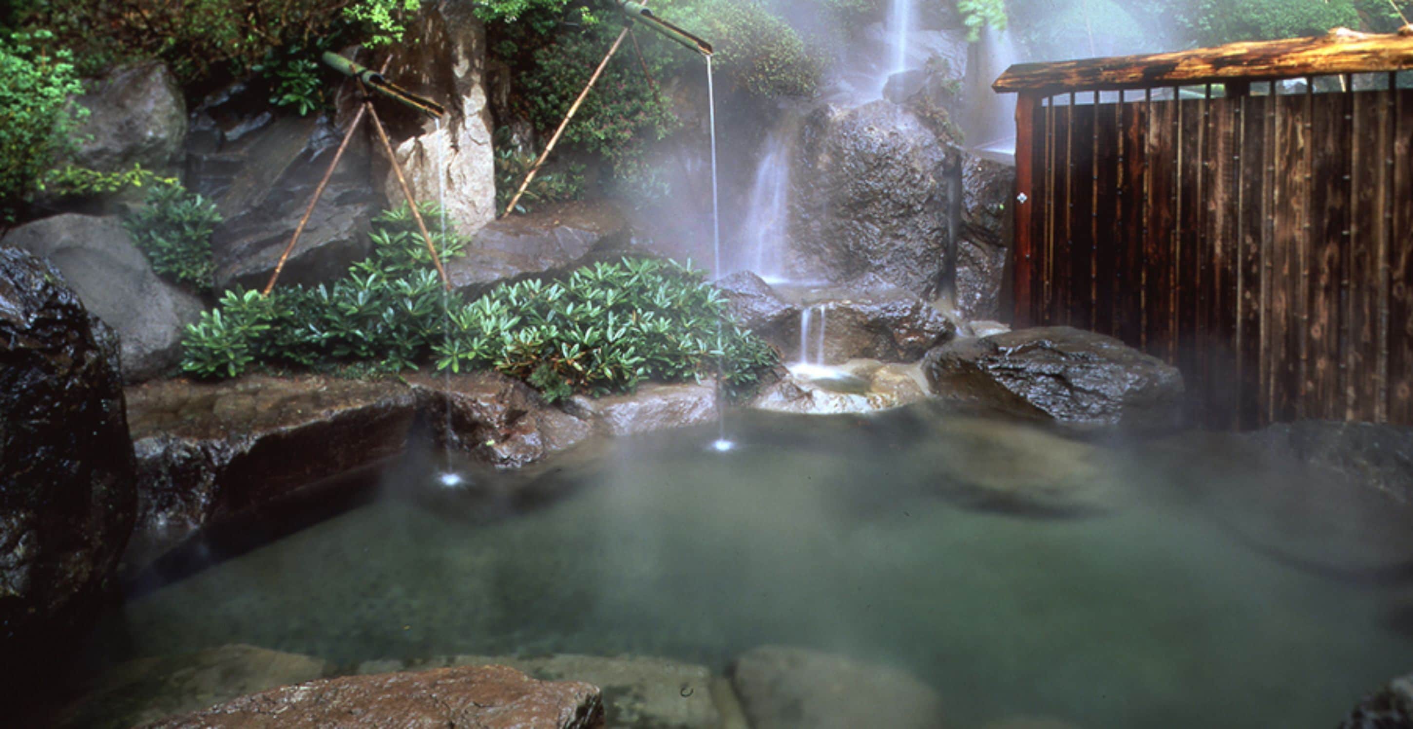 A natural hot spring with a wooden viewing platform, surrounded by rocks and greenery.