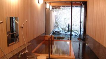 Interior de madera con bañera de hidromasaje con cubierta de vidrio, suelo de madera y vistas a una zona exterior rocosa.