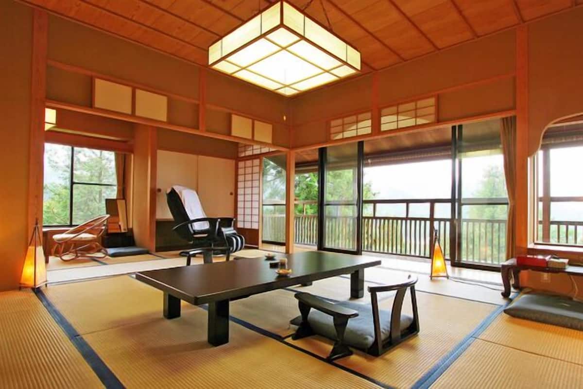 Een traditionele kamer met tatami matten, een tatami tafel, een zwarte stoel en uitzicht op het buitenleven door grote ramen.