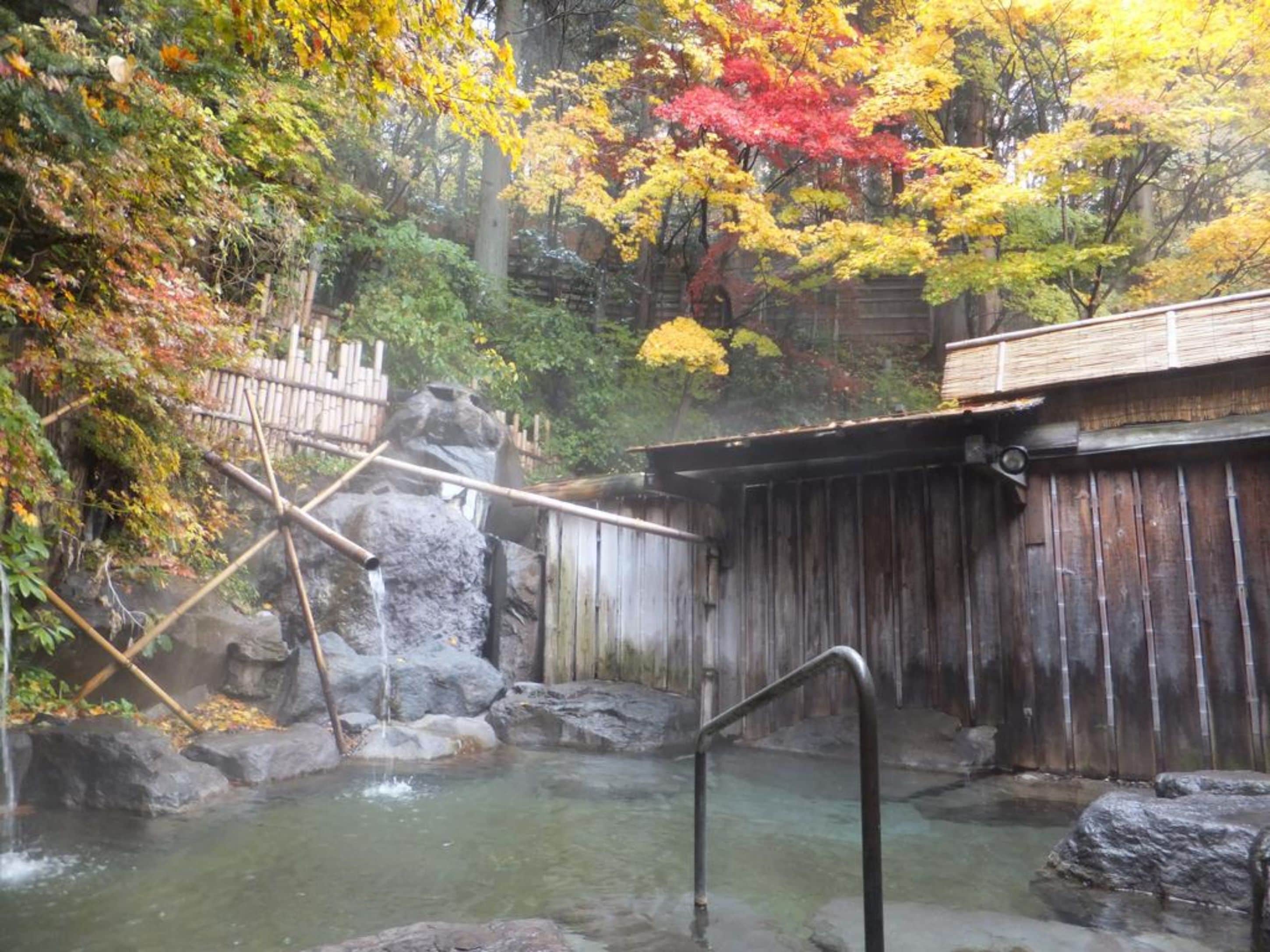 A traditional Japanese onsen with a wooden bathhouse, a small waterfall, and a rocky landscape.