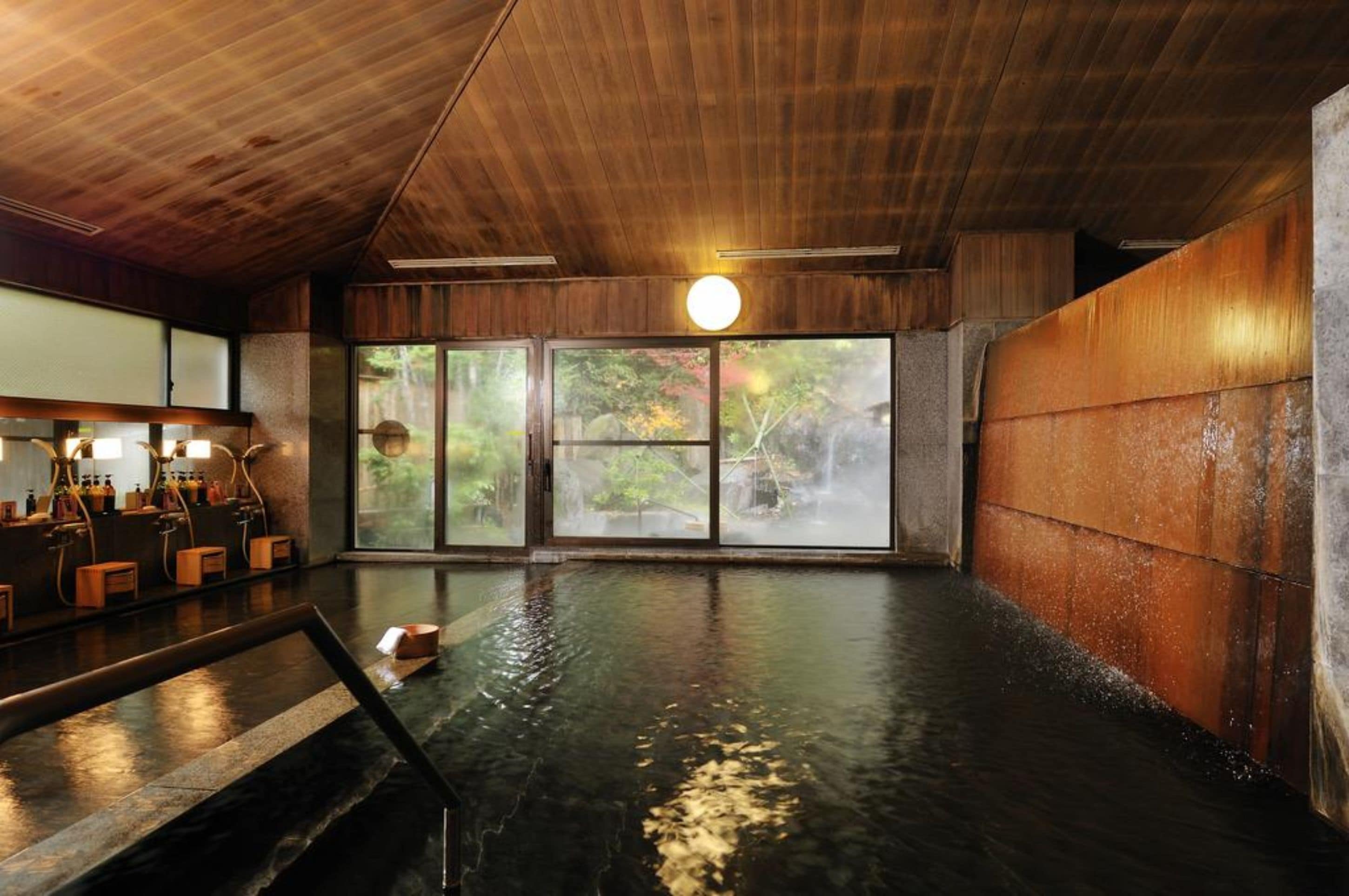 A traditional Japanese onsen with wooden interiors, a stone bath, and a view of nature through large windows.
