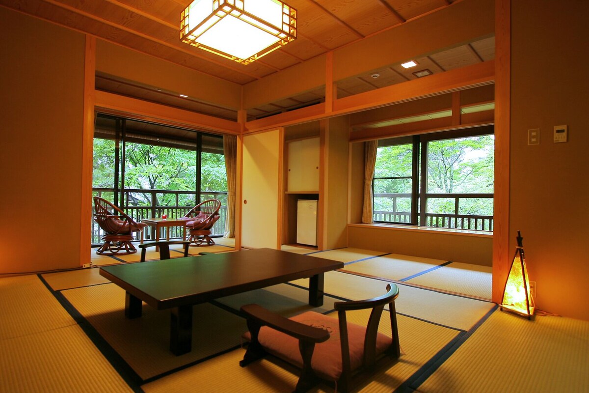 A traditional Japanese room with tatami flooring, a low table, chairs, and large windows with a view of greenery.