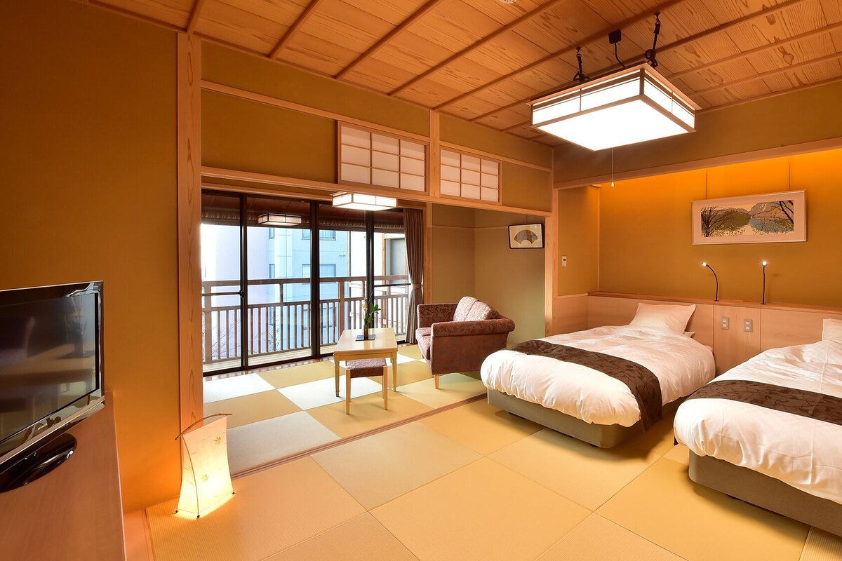 A hotel room with two beds, a TV, a desk, and a view of the outside.