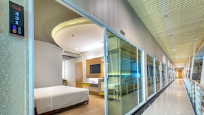 Quarto de hotel com uma cama, escrivaninha, televisão e parede de vidro.