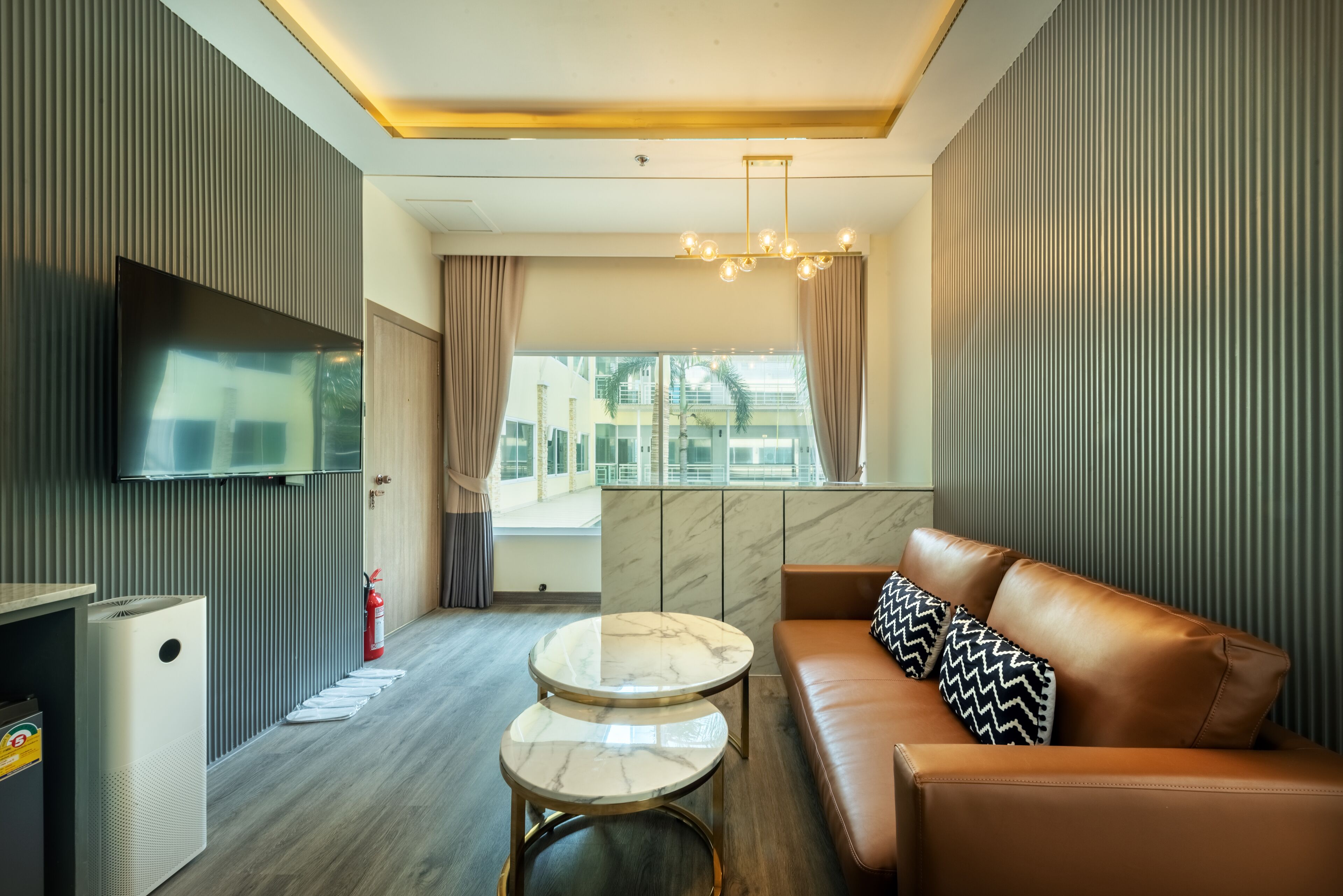 Une chambre d’hôtel moderne dotée d’une grande télévision à écran plat, d’un canapé agrémenté de coussins à motifs, d’une table basse en marbre et offrant une vue sur la ville grâce à de grandes fenêtres.