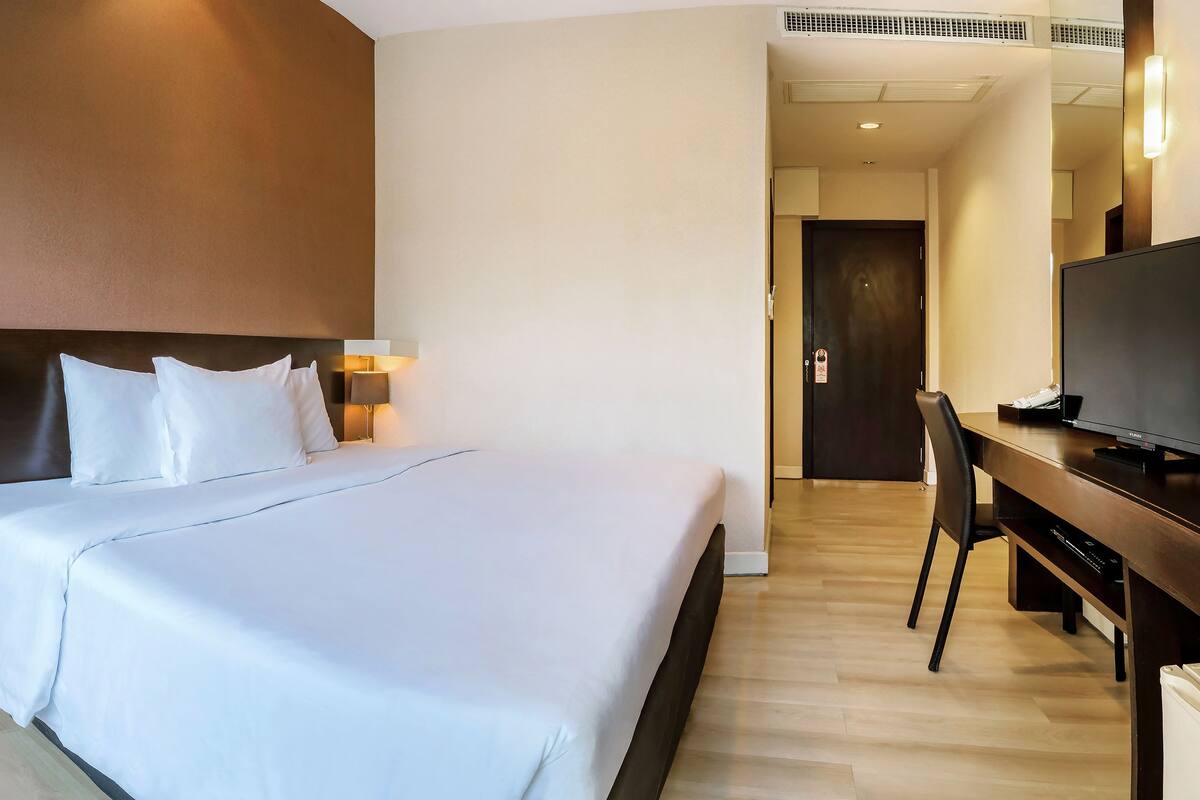 Una habitación de hotel con una cama grande, lámparas de noche, un escritorio con televisor y una puerta que da a un pasillo.