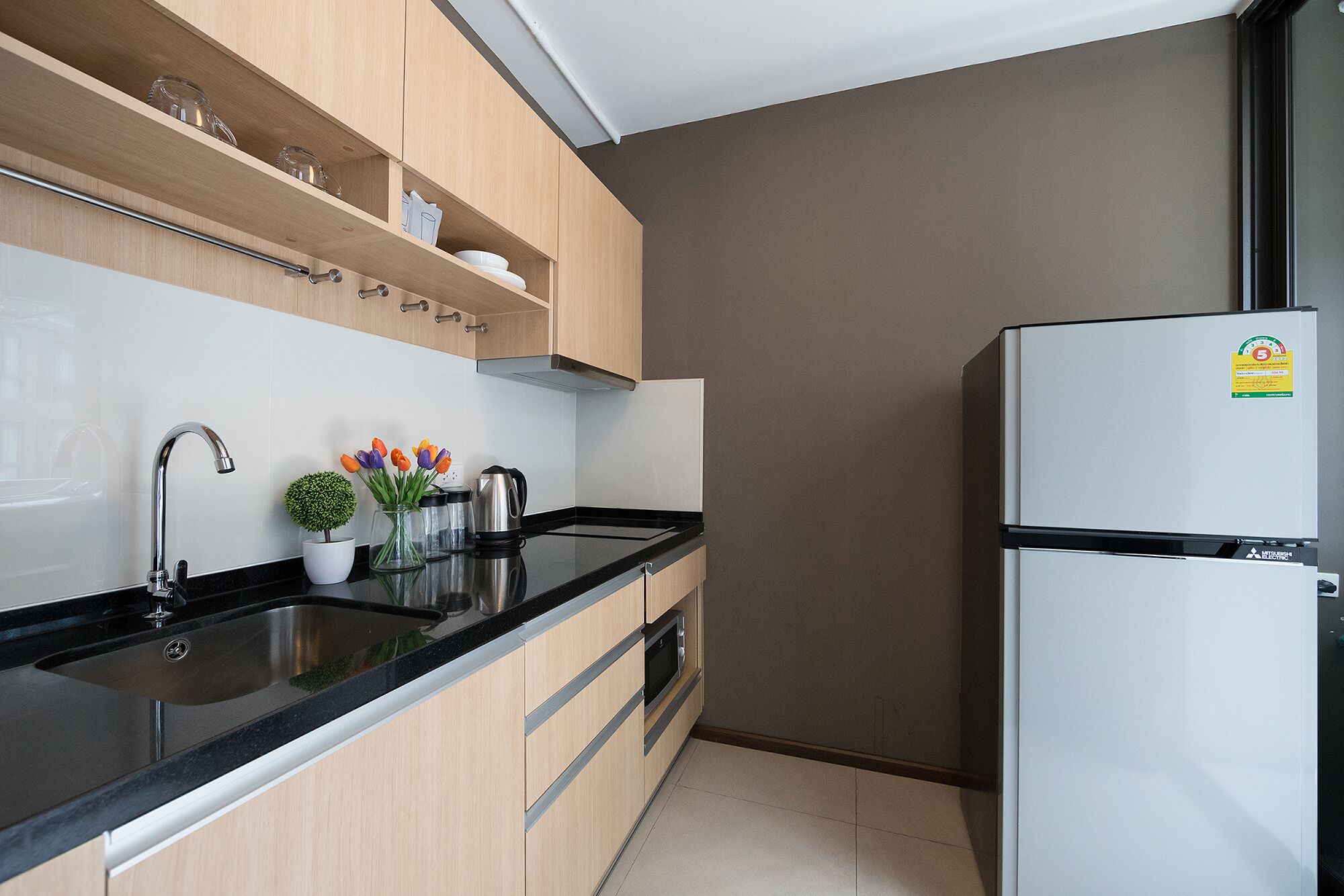 A modern kitchen with a black countertop, stainless steel sink, wooden cabinets, a white refrigerator, and a small potted plant with flowers.