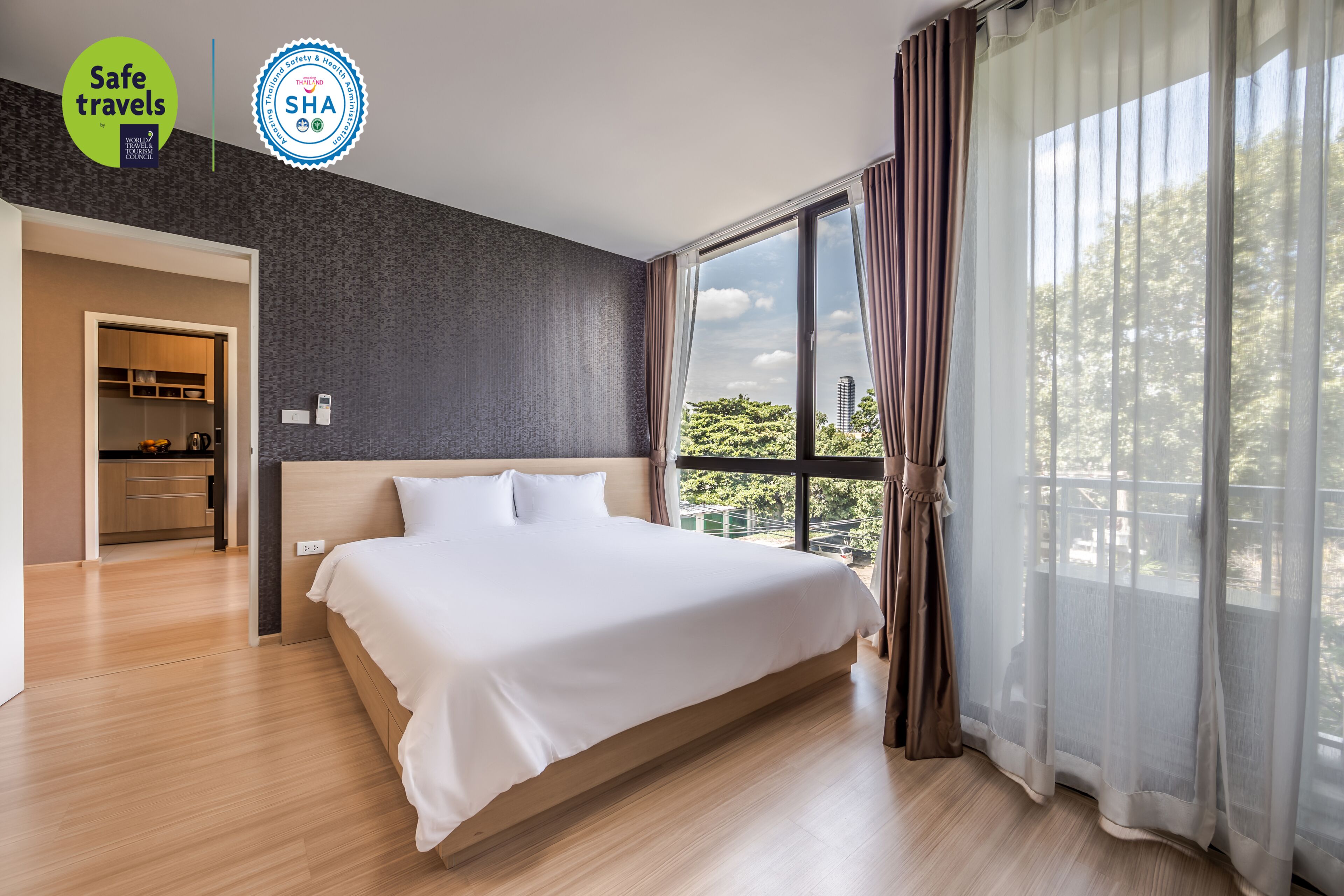 Ein modernes Hotelzimmer mit einem großen Bett, Blick auf Grünflächen und ein Gebäude durch transparente Vorhänge, sowie eine kleine Küche im Hintergrund.