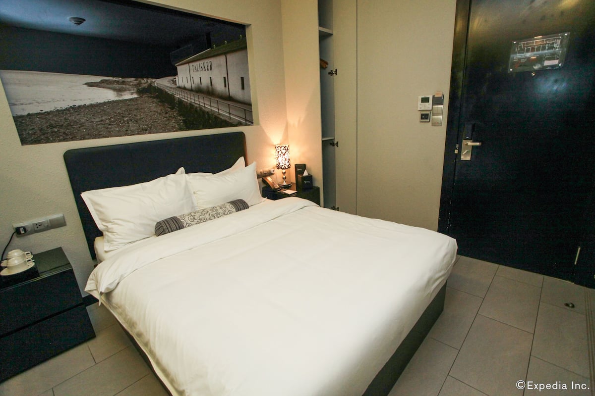 Habitación de hotel con una cama bien hecha, una mesita de noche con lámpara, una fotografía en la pared y una puerta con cerradura.