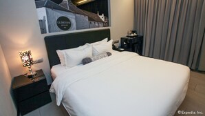 Ett hotellrum med en stor säng, ett nattduksbord med en lampa, en väggmonterad TV och utsikt över en byggnad genom ett fönster.