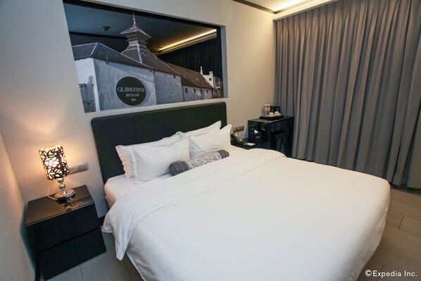 A hotel room with a large bed, a nightstand with a lamp, a wall-mounted TV, and a view of a building through a window.