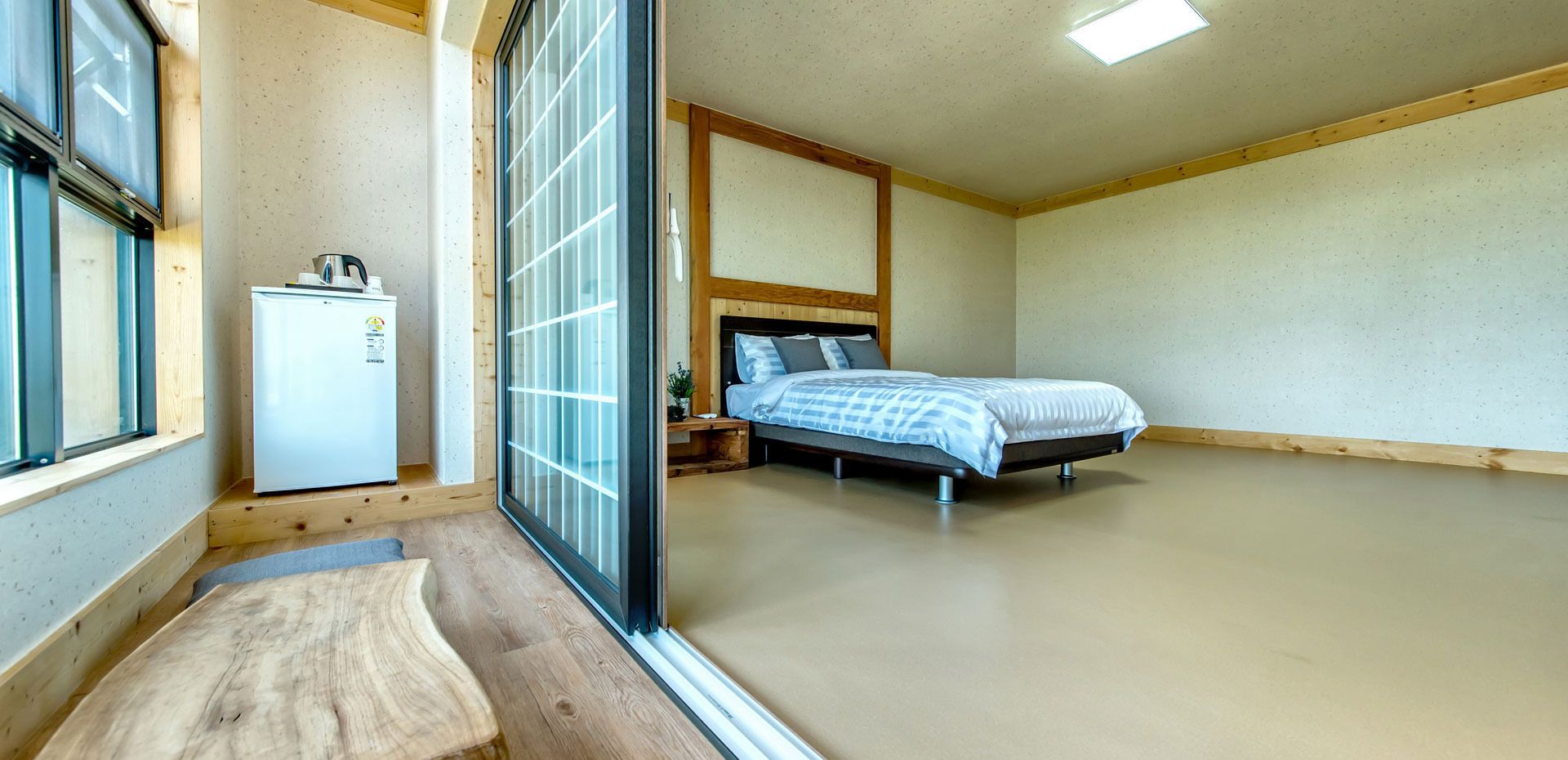 A bedroom with a bed, a small bedside table, a nightstand, a window, and a refrigerator.