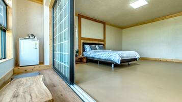 A bedroom with a bed, a small bedside table, a nightstand, a window, and a refrigerator.