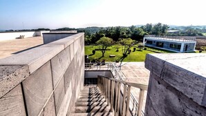 Un edificio moderno con fachada de concreto, una escalera que lleva a una zona en la azotea y vistas de un jardín ajardinado con árboles y un paisaje distante.