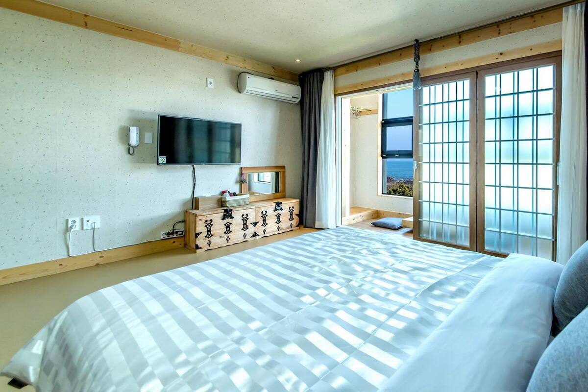 A hotel room with a large bed, a wooden dresser, a flat-screen TV, and a view of the sea through a shoji screen.