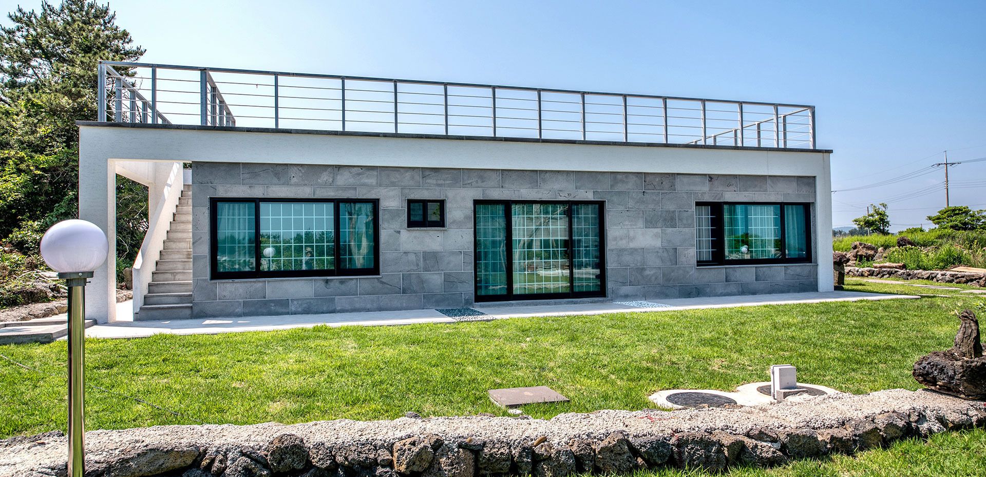 Ein modernes, zweistöckiges Gebäude mit großen Glasfenstern, einem Metallgeländer im oberen Stockwerk und einer Steinmauer im Vordergrund.