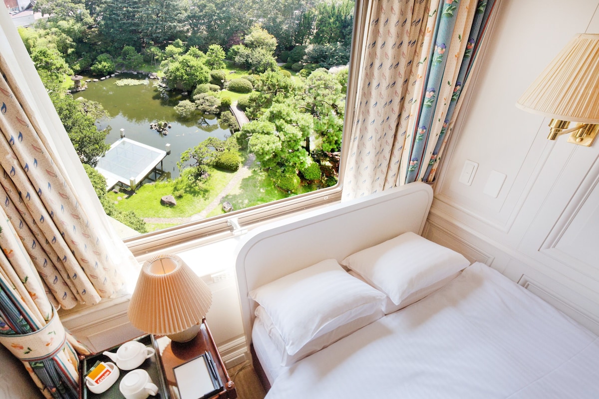 Habitación de hotel con cama, mesita de noche con lámpara, bandeja con té y tetera, y vista a un jardín con estanque.