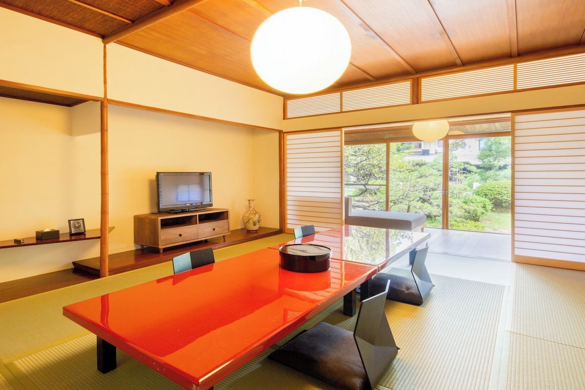 Una habitación tradicional japonesa con suelo de tatami, una mesa baja, televisión y vistas a la vegetación exterior.