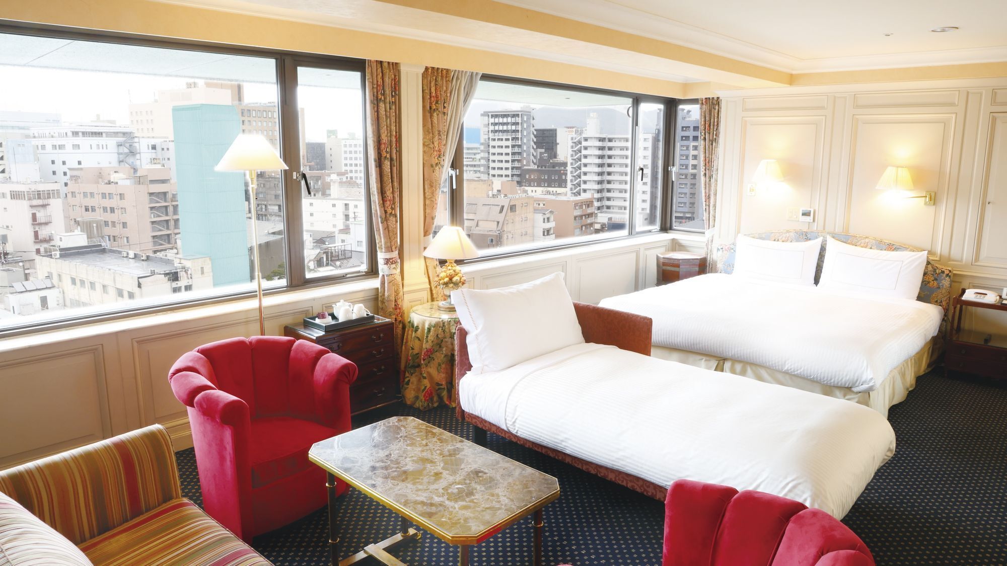 A hotel room with two beds, a red armchair, a small table, and a view of the cityscape.