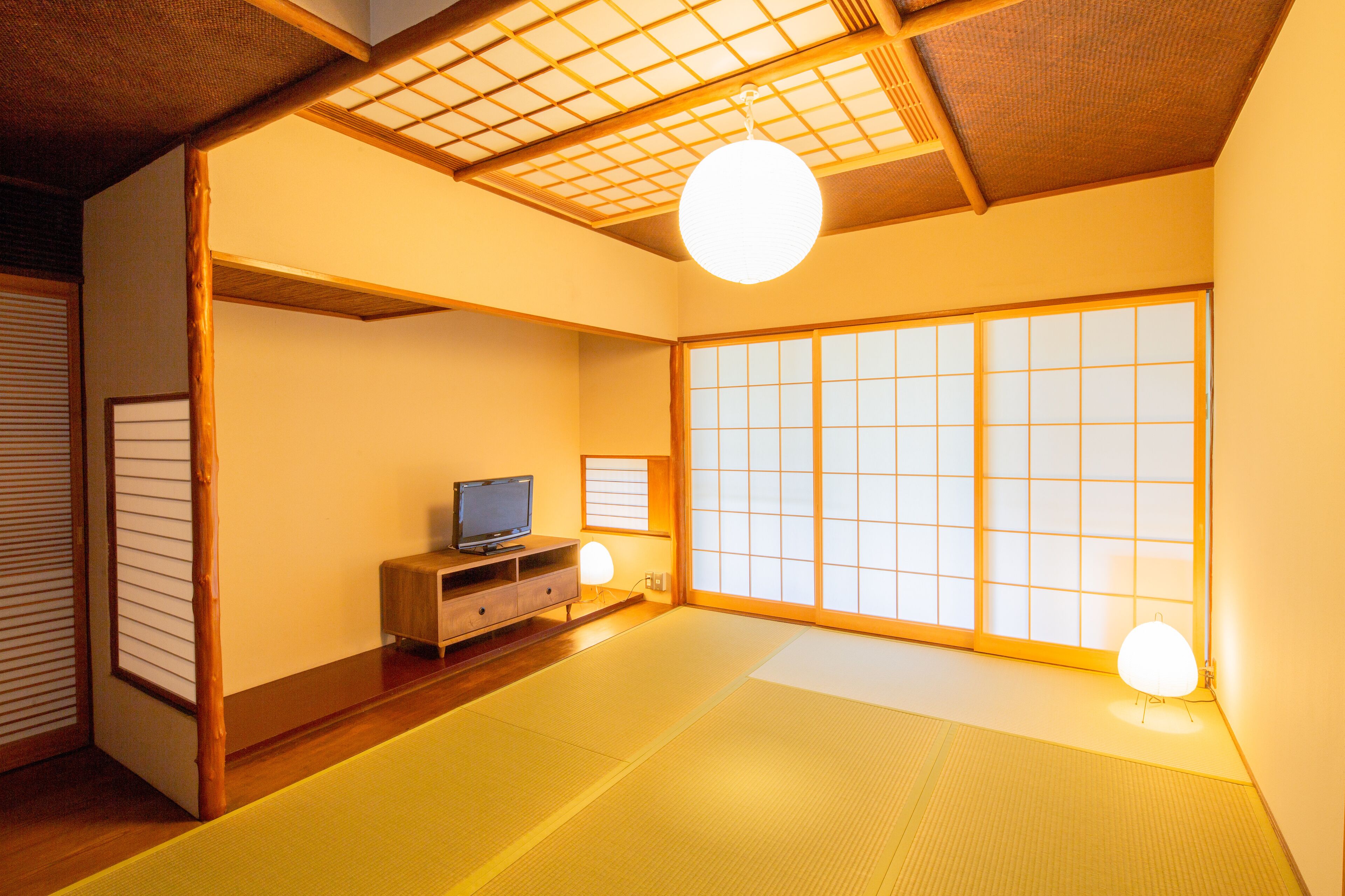 A traditional Japanese room with tatami flooring, a television on a wooden stand, sliding shoji doors, and a hanging light fixture.