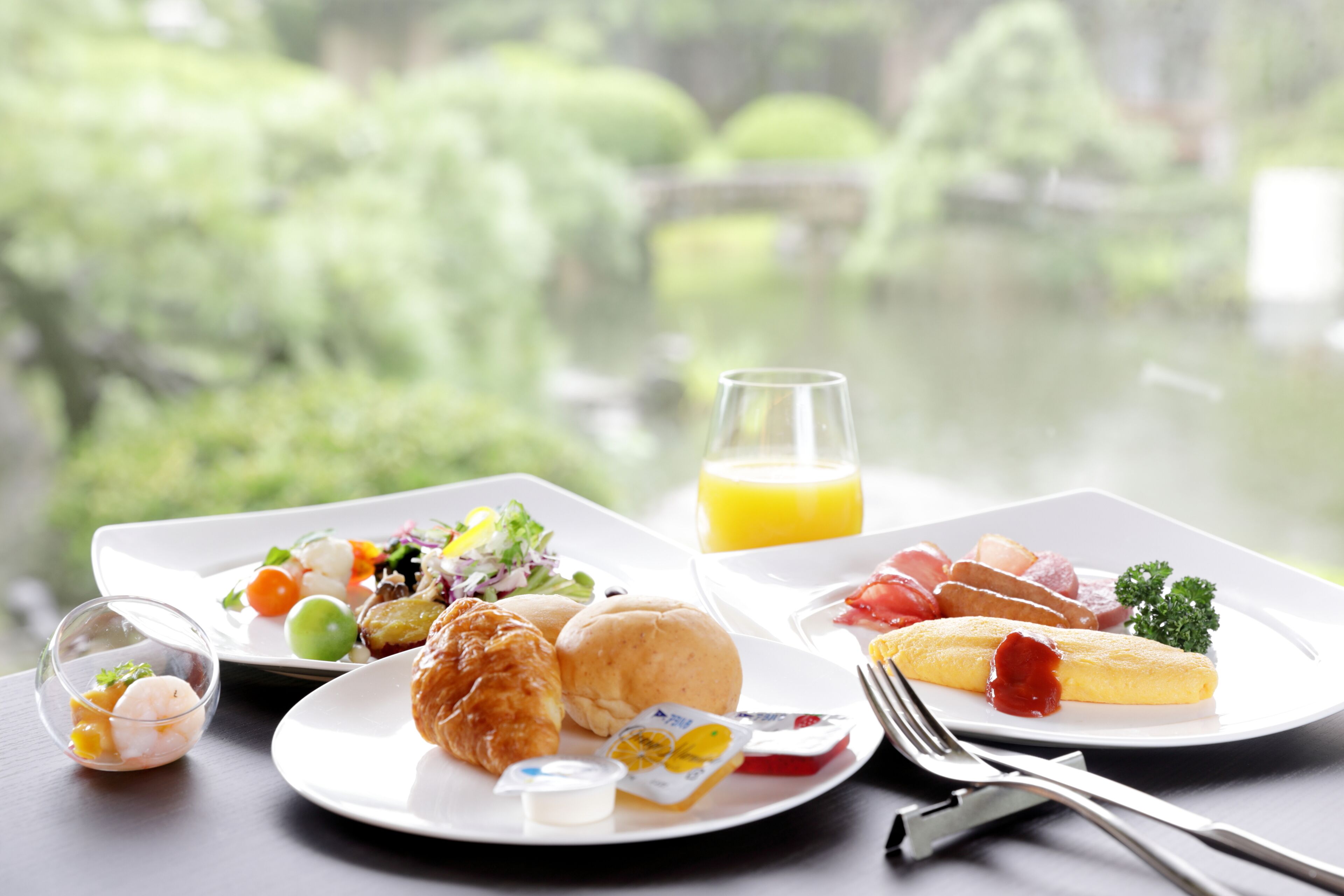 A breakfast setting with croissants, eggs, and a glass of orange juice on a table with a view of greenery.