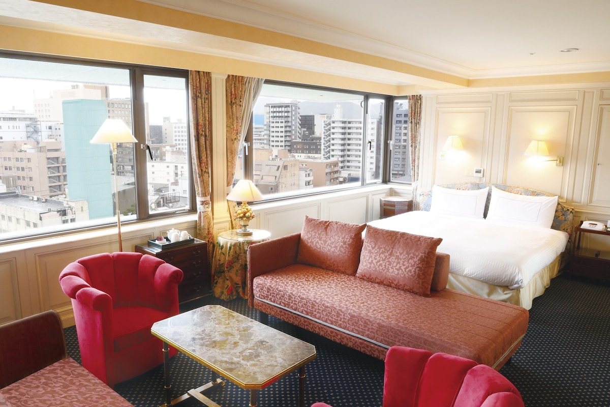 Habitación de hotel con una cama grande, un sofá rojo, una mesita y vistas de la ciudad a través de ventanas amplias.