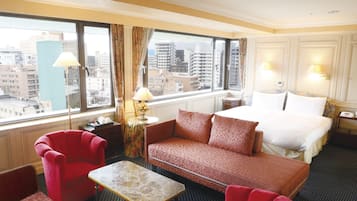 Une chambre d’hôtel avec un grand lit, un canapé rouge, une petite table et une vue sur la ville grâce à de grandes fenêtres.