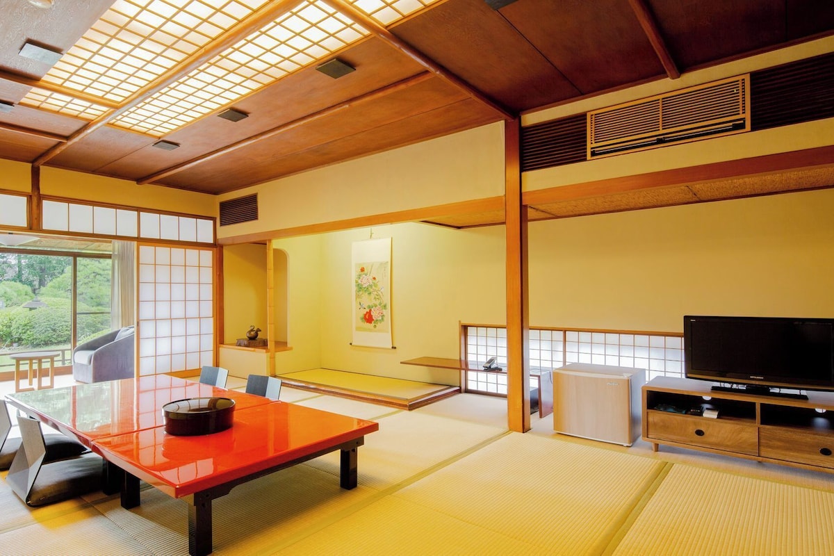 Una habitación tradicional japonesa con suelo de tatami, una mesa baja, un sofá, un televisor y una puerta corrediza que da a un jardín.