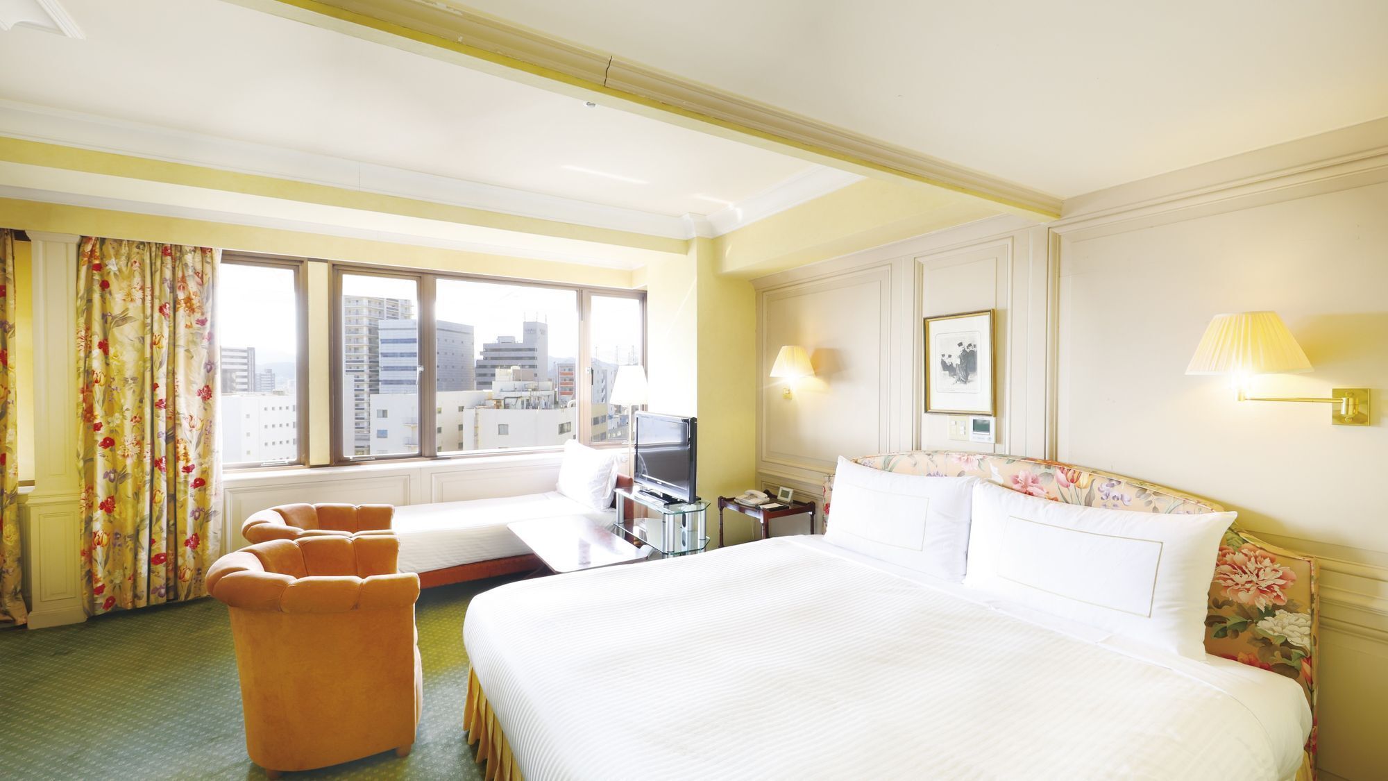 A hotel room with a large bed, a desk with a television, a sofa, and a view of the cityscape through large windows.
